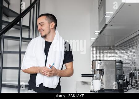 Ein junger Mann geht morgens zur Arbeit. Bürsten der Zähne in der Nähe der Kaffeemaschine beim Warten auf eine Tasse Kaffee. Stockfoto