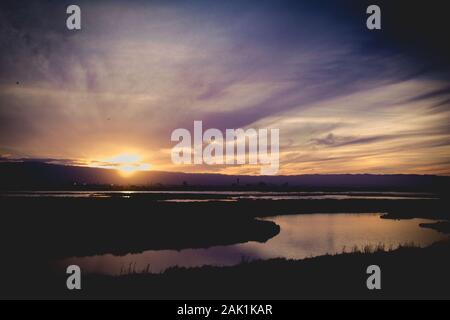 Die Sonne geht hinter den Bergen unter, hinter dem sich schlänfenden Fluss an einem wolkigen Wintertag, der von unten beleuchtet wird. Lila Dunst, eine Spiegelung des Himmels. Stockfoto