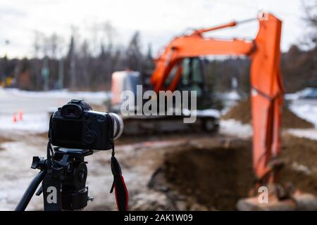 Eine selektive Fokus Ansicht einer Kamera auf einem Stativ fotografieren ein Bagger arbeiten auf einer Baustelle, sehen unscharf im Hintergrund mit Kopie Raum Stockfoto