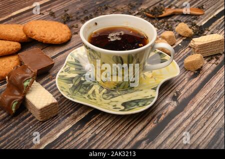 Eine Tasse schwarzer Tee, Teeblätter, Stücke von braunem Zucker und Haferflocken Kekse, Waffeln, Stücke Schokolade auf einem hölzernen Hintergrund. Nahaufnahme Stockfoto