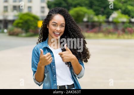 Brasilianische Mädchen mit lockigem Haar und Klammern zeigen beide Daumen nach oben Outdoor im Sommer in der Stadt Stockfoto