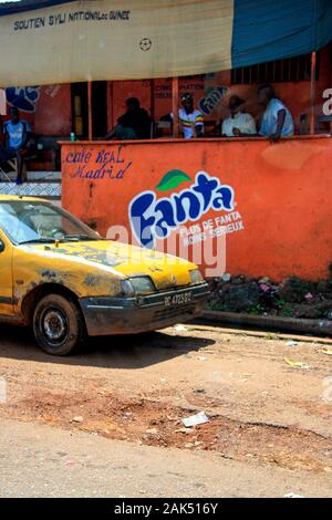 Blick auf ein altes gelbes Auto, das auf der Seite der Feldstraße vor dem Café Real Madrid in Conakry, Guinea, geparkt ist Stockfoto