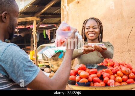 Afrikanisches Mädchen Verkauf von Tomaten in einem lokalen afrikanischen Markt zu einem männlichen Kunden Lächeln beim Empfang der Zahlung Stockfoto