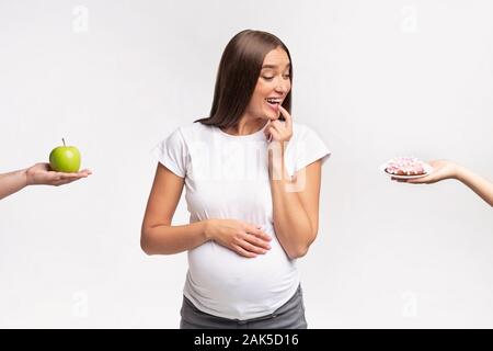 Schwangere Frau Diät. Aufgeregt werdende Dame der Wahl zwischen Donut und Apfel Obst stehen auf weißen Studio Hintergrund Stockfoto