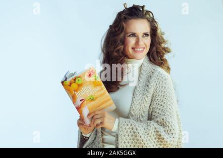 Lächelnde junge Frau mit langen gewelltes Haar in Pullover und Strickjacke mit gesunder Ernährung Buch bei copy Speicherplatz auf Winter hellblau hinterlegt. Stockfoto