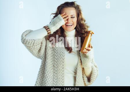 Lächelnde junge hausfrau mit langen gewelltes Haar in Pullover und Strickjacke holding Self Tanning Lotion isoliert auf Winter Hellblau. Stockfoto