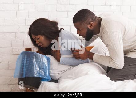 Morgendliche Übelkeit. Junge schwangere afrikanische Frau sitzt auf dem Bett, Papierkorb, Gefühl der Übelkeit während der Schwangerschaft, ihr Mann ihr tröstend Stockfoto