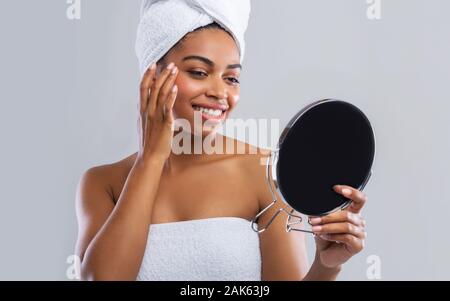 Hübsche schwarze Frau in ein Handtuch eingewickelt ihr Gesicht im Spiegel prüfen nach Dusche über grauer Hintergrund, Hautpflege Konzept Stockfoto