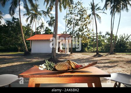 Luxuriöse Unterkunft in tropischen Ziel. Obst in der Schüssel auf hölzernen Tisch gegen Bungalow unter Kokospalmen. Stockfoto