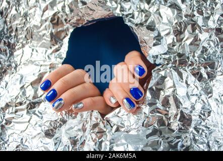 Die junge Frau Hände mit schönen Maniküre blau auf blauem Hintergrund mit metallisch glänzendes Papier. 2020 color Trend. Stockfoto