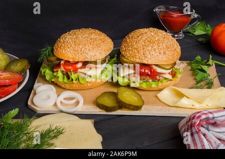 Fertige Hamburger mit Zutaten auf einem Holzbrett auf schwarzem Holz- Hintergrund. Für Werbung, Menü. Von der Seite. Stockfoto