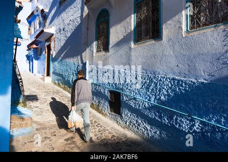 Mann, der Plastiktüten mit Orangen und Bananen in der sonnigen Straße Medina von Chefchaouen (auch Chaouen genannt), Marokko, trägt Stockfoto