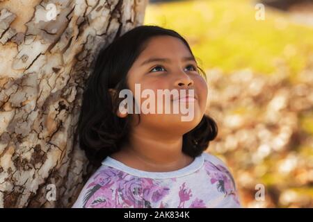 Ein latino Mädchen mit einem sanften Lächeln, lehnte sich an einen Baum und blickte in den Himmel mit einem Gefühl der Hoffnung. Stockfoto