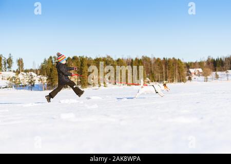 Winter in Finnland Konzept mit Jungen, die nach dem Hund auf dem Eis des zugefrorenen See mit Dorf im Hintergrund Stockfoto