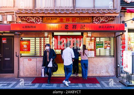 Chinatown Nankin-Machi in Motomachi, Kobe, Japan. 3 Leute in der Warteschlange außerhalb Eingang mit noren Vorhänge eines chinesischen Knödel mitnehmen. Am Abend Stockfoto