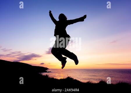Silhouette einer Frau vor Freude hüpfend auf küstennahen Klippen bei Sonnenuntergang Stockfoto