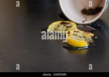 Dunkle Tisch mit einer Tasse Kaffee verschüttet. Zitronenscheiben und Teeblätter mit Tee Reste auf dem Tisch. Von der Seite. Selektive konzentrieren. Augenhöhe schießen Stockfoto