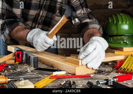 Close-up. Tischler mit seinen Händen durch Handschuhe geschützt, mit Hammer und Nägel behebt ein Holzbrett. Bauwirtschaft, die es selbst tun. Holz- arbeiten Stockfoto