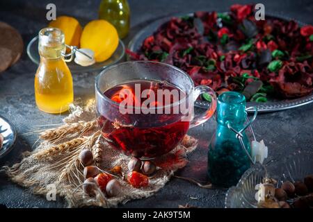 Red Hot Hibiskus Tee in einen Glaskrug auf einem Holztisch unter Rosenblättern und trockenen Tee Creme mit metallischen Herz Stockfoto