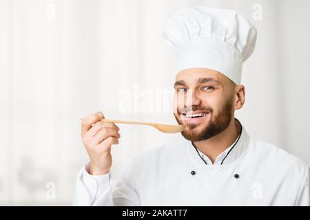 Happy Chef Mann Tastingnahrung Holding Löffel lächelnd an Kamera stehend gegen Fenster in Küche Indoor. Stockfoto