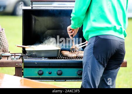 Ein Mann kochen Würstchen auf einem offenen Gas bbq, gibt es auch ein Fach der Zwiebeln darauf warten bedient zu werden. Stockfoto
