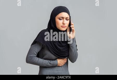 Notruf. Sick muslimischen Frau im hijab mit akuten Abdomen Magenschmerzen, um Hilfe für den Arzt, über grauer Hintergrund stehend Stockfoto