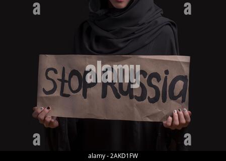 Muslimisch-arabischen Mädchen. Russland Aggressor. Arabische Trauriges Mädchen in schwarzen Abaya und schwarzer Hintergrund mit Plakat mit Plakat stoppen Russland, keinen Krieg. Stockfoto