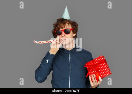 Feier Konzept. Teenager Junge in Geburtstag Kappe und Sonnenbrille isoliert auf Grau mit vorhanden Blasen party Horn lächelte zuversichtlich stehend Stockfoto