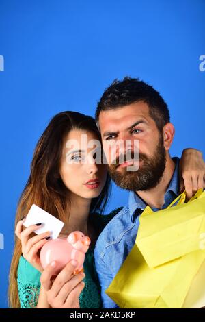 Kerl mit Bart und Mädchen mit ernsten Gesichtern einkaufen. Shopping und Freizeit Konzept. Paar in der Liebe hält Einkaufstaschen auf blauem Hintergrund. Mann mit Bart hält Kreditkarte und Geld. Stockfoto