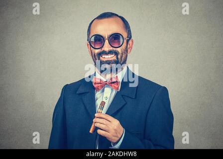 Lächelnd zuversichtlich, bärtigen Mann trägt runde Sonnenbrille, hält kubanische Zigarre. Portrait von stilvollen, lustiger Kerl suchen Sie Kamera. Moderne Kleidung dunkler Anzug s Stockfoto