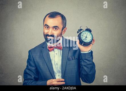 Closeup Portrait stattlich, erfreut nach bärtigen Mann in elegante Jacke mit roter Schleife binden Holding übersicht Wecker in der Hand, grauen Hintergrund. Stockfoto