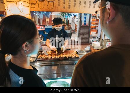 Zwei Kunden beobachten, wie ein Verkäufer gegrillte King Auster Pilze auf dem Shi Lin Night Market, Taipei, Taiwan, zubereitet Stockfoto