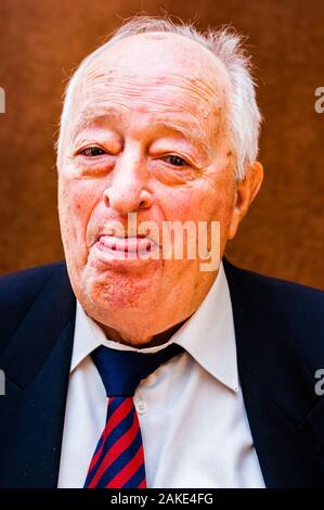 Portrait von positiven weißen älteren Mann mit dunkler Anzug, weißes Hemd und Krawatte gestreift blau rot seine Zunge, mit einem Auge auf hellen braunen Hintergrund Stockfoto