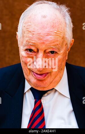 Portrait von Weißen älteren Mann mit dunkler Anzug, weißes Hemd und Krawatte gestreift blau rot heimlich mit der Kamera auf der Suche nach hellen braunen Hintergrund Stockfoto