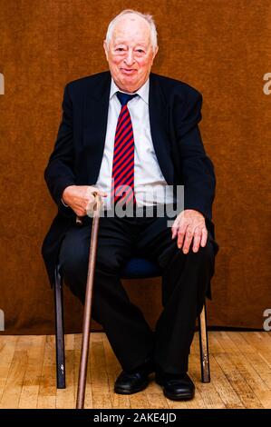 Portrait von Happy white älterer Mann mit dunkler Anzug, weißes Hemd und Krawatte gestreift Blau Rot lächelnd saß auf einem Stuhl und halten die Hand auf wenige Stic Stockfoto