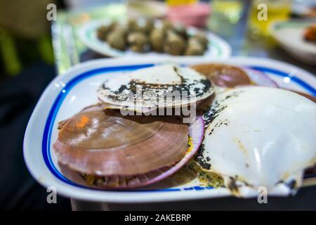 Bangkok/Thailand-December 2019: Gegrillte Jakobsmuscheln auf einem Teller im Vordergrund und gekochte Herzmuscheln im Hintergrund auf einem Tisch in der Nacht in Chinatown Stockfoto