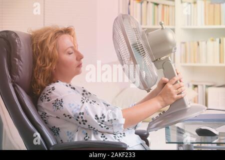 Frau leidet unter der Hitze, während der Arbeit im Büro und versucht durch den Lüfter zu kühlen Stockfoto