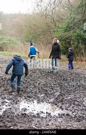 Mutter/Mutter/Mutter/Mutter, die mit ihren drei Kindern an einem Wintertag auf einem schlammigen Weg durch Wälder auf West End Common, Esher, Surrey durch den Schlamm spazieren. GROSSBRITANNIEN. (115) Stockfoto