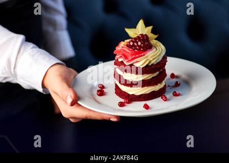 Köstliche Kuchen aus rotem Samt mit Schlagsahne, Granatapfel und karambolen auf weiße Platte in weibliche Hände eingerichtet. Stockfoto