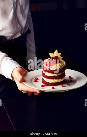 Köstliche Kuchen aus rotem Samt mit Schlagsahne, Granatapfel und karambolen auf weiße Platte in weibliche Hände eingerichtet. Stockfoto