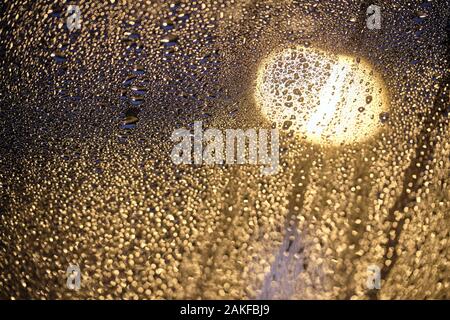 Moody helle Foto von schillernden Regentropfen auf Glas vor eine Straßenlaterne in der Nacht. An einem regnerischen Tag in Deutschland im Januar gesehen auf einem Dach, Fenster Stockfoto