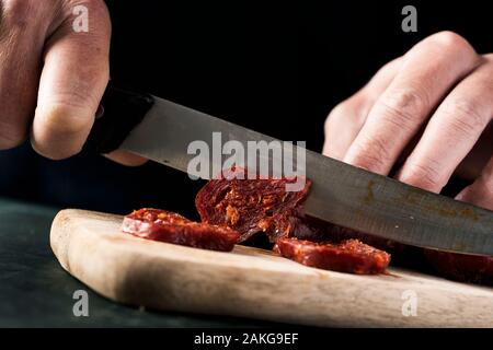 Nahaufnahme von einem Mann schneiden einige Scheiben Chorizo, einem ausgehärteten Wurst aus Schweinefleisch, die typisch für Spanien, mit einem Küchenmesser auf einer hölzernen Schneidebrett Stockfoto