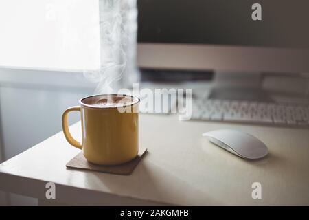 Eine gelbe Tasse Kaffee steht auf dem Schreibtisch im sonnigen Morgen abend Licht, neben dem Monitor und der Tastatur Stockfoto