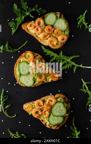 Leckere Toasts mit gebratenen Garnelen, Avocado und Gurke auf schwarzen Hintergrund. Flach Stockfoto