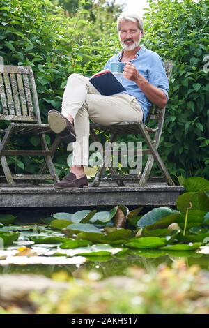 Portrait Des Reifen Mannes, Der Sich Im Buch "Garden Reading Book On Jetty By Lake" Entspannen Kann Stockfoto