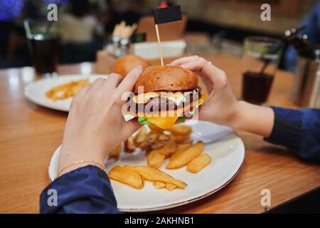 Saftiger Käseburger in Mädchen-Händen. Naturbelassener Rinderburger mit Doppelkäse serviert mit Kartoffeldippern. Kalorienreiches Abendessen im Café. Stockfoto