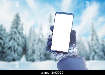 Telefon mockup in Hand mit Handschuhen. Winter, kalte Zeit mit viel Schnee im Gebirge. Isolierte Bildschirm für Mockup, App. Verschneite Bäume in backgrou Stockfoto