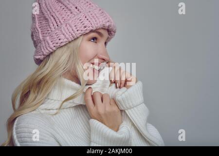 Hübsche junge blonde Frau in der kammgarn winter Outfit kuscheln in die Wärme ihres rollkragen pullover mit einem glücklichen Lächeln, während ein rosa gestrickt tragen kann Stockfoto