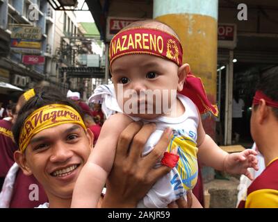 Ein Baby mit einem nazaräer Stirnband, heben sie von seinem Vater während der Veranstaltung. Filipinos erfassen am Quiapo in Manila für das Fest des Schwarzen Nazareners, eine lebensgroße Statue eines leidenden Jesus Christus, der vom Kreuz gefallen. Katholische Gläubige tun ' pakaridad" einen Weg geben oder das gemeinsame Essen mit anderen und eine lange Prozession der Statue. Stockfoto