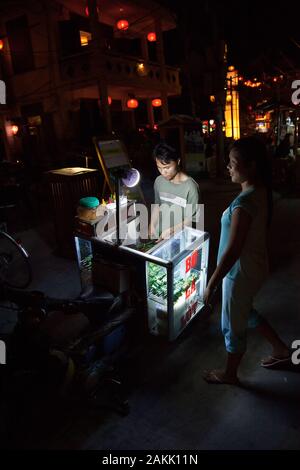 Asiatischer Straßenhändler mit Lebensmittel-Stall in der Nacht. Trolley auf Rädern mit Imbiss, verkauft Straßennahrung in Vietnam. Stockfoto